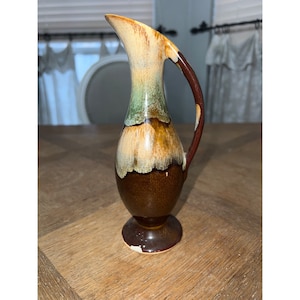 Könnte beinhalten: Ein kleiner, brauner Keramik Krug mit grüner und gelber Glasur. Der Krug hat einen runden Körper und einen kleinen, ausgestellten Boden. Er hat einen Griff an der Seite.