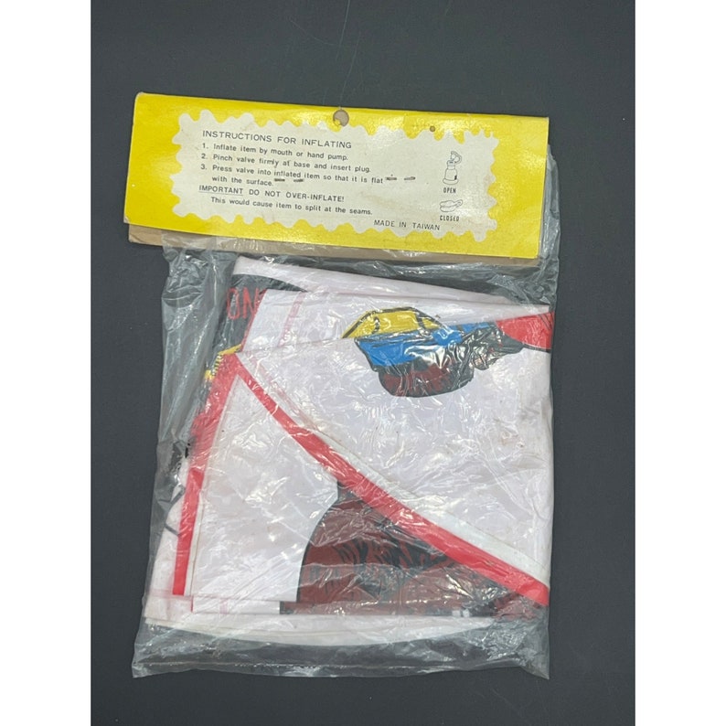 May include: A white inflatable pool float with a red and black cartoon car design. The float has a yellow car with a blue windshield and a red driver. The packaging includes instructions for inflating the float. The instructions say to inflate the item by mouth or hand pump, pinch the valve firmly at the base and insert the plug, and press the valve into the inflated item so that it is flat with the surface. The instructions also say to not over-inflate the item, as this would cause it to split at the seams. The packaging is made in Taiwan.