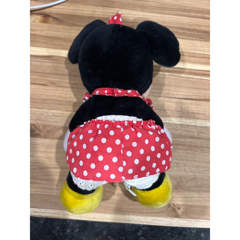 Pu&ograve; includere: Un giocattolo di peluche nero e rosso di Minnie Mouse con un vestito a pois rossi. Il giocattolo &egrave; seduto su una superficie di legno.