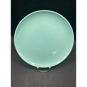 May include: A round, pale teal ceramic plate. The plate has a smooth, glossy finish and is displayed on a gold-coloured metal stand. The plate is a solid colour with no visible patterns or designs.