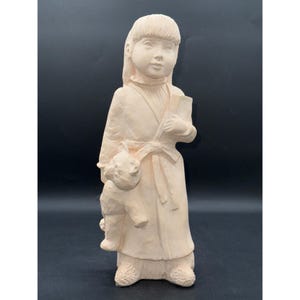 May include: A beige ceramic figurine of a young person holding a book and a small animal. The figure is wearing a long coat with a tied belt and has long hair. The figurine is standing on a textured base.