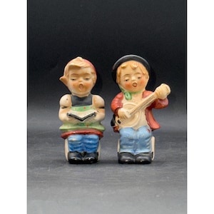 May include: Two vintage ceramic figurines of children. One is singing from a book, wearing a red cap, black top, green collar, and blue pants. The other plays a banjo, wearing a black hat, red jacket, green scarf, and blue pants. Both have black shoes.