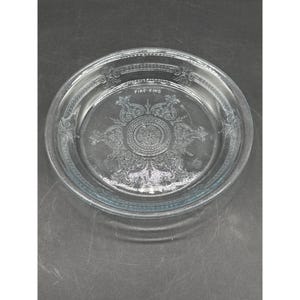 Peut inclure: Plat à tarte Fire-King en verre transparent avec un motif en relief orné. L'assiette présente un médaillon central et une bordure décorative. Le texte "FIRE-KING" est visible.