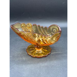 Könnte beinhalten: Bernsteinfarbene Vintage-Glasschale mit Sockel. Die Schale hat eine bootsartige Form mit einem gewellten Rand und komplizierten Stern- und Wirbelmustern. Der Sockel ist rund mit einem Blumendesign.