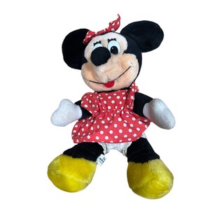 Pu&ograve; includere: Un giocattolo di peluche di Minnie Mouse che indossa un vestito rosso con pois bianchi e un fiocco rosso. Il giocattolo ha occhi neri, un naso rosso e un sorriso giallo. Il giocattolo &egrave; seduto su uno sfondo bianco.