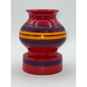 Pode incluir: Um vaso de cerâmica vermelho com uma base larga e um pescoço estreito. O vaso é decorado com listras horizontais amarelas, roxas e vermelhas.