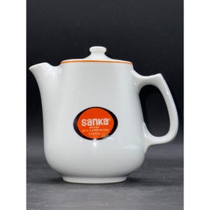 May include: White ceramic Sanka brand coffee pot with a lid and handle. The pot has an orange rim and an oval logo that reads "sanka" and "97% caffeine free coffee".