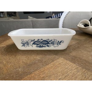May include: A rectangular white baking dish with a blue floral design. The dish has a slightly curved rim and is sitting on a wooden table. The blue design features stylized leaves and berries.