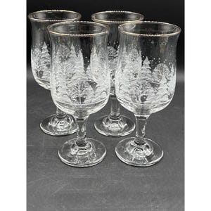 May include: A set of four clear wine glasses, each featuring an etched winter forest scene. The glasses have gold rims and a round base. The forest design includes trees and falling snow.