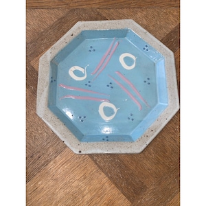 Puede incluir: Plato octogonal de cerámica con un esmalte azul claro y un diseño geométrico blanco y rosa. El plato tiene un borde blanco moteado.