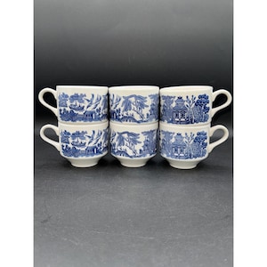 May include: Six white teacups with blue transferware designs are stacked in two rows. Each cup features a different scenic pattern, including pagodas, trees, and boats. The cups have curved handles and a classic, vintage aesthetic.