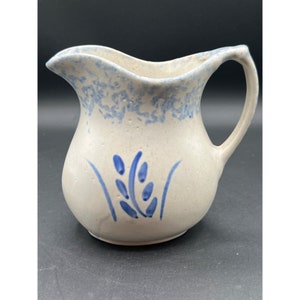 Puede incluir: Una jarra de cerámica blanca con un diseño floral azul. La jarra tiene un esmalte moteado y un asa.