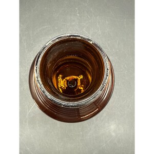 May include: An overhead view of a brown glass bottle. The bottle has a wide, circular opening and a ridged exterior. The interior of the bottle is visible, with a light source reflecting off the glass.