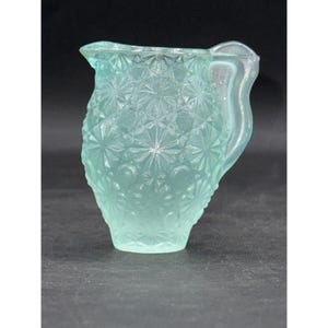 May include: A vintage, light blue glass pitcher with a starburst pattern. The pitcher has a handle and a spout for pouring. The glass has a frosted appearance, and the pitcher is likely used for serving beverages.