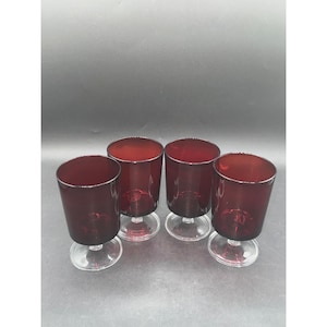 May include: A set of four matching drinking glasses featuring deep red, translucent bowls and clear glass stems with circular bases. Each glass has a smooth, cylindrical shape and a consistent, vibrant ruby color throughout the glass body.