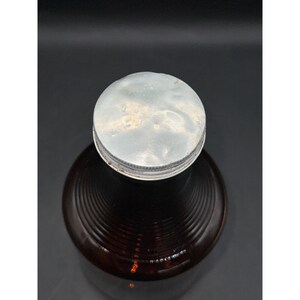 May include: A vintage brown glass bottle with a silver metal lid. The bottle has a ribbed design and is filled with a dark liquid. The lid shows signs of age and wear, adding to its antique appearance.