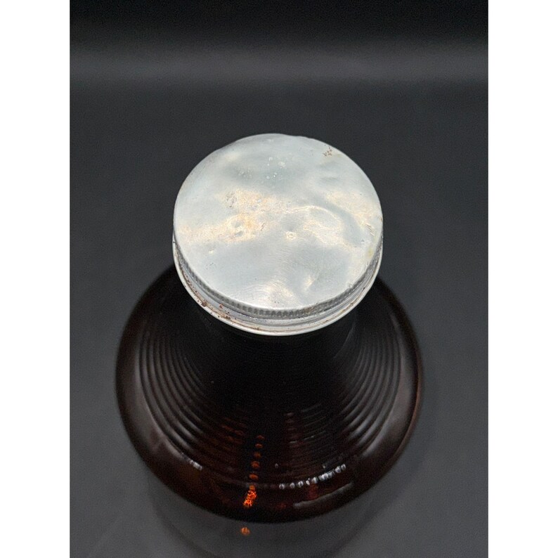 May include: A vintage brown glass bottle with a silver metal lid. The bottle has a ribbed design and is filled with a dark liquid. The lid shows signs of age and wear, adding to its antique appearance.