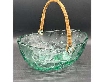May include: A clear green glass basket with a woven rattan handle. The basket has a decorative pattern of grapes and leaves. The handle is attached to the basket with clear glass beads. The basket is designed for serving fruit or as a decorative piece.