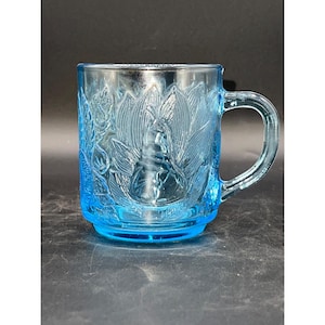May include: A light blue glass mug with a floral design. The mug has a handle and is made of clear glass with a frosted design.