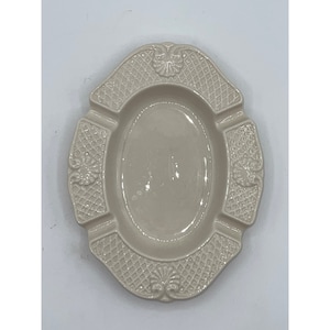 May include: A white ceramic ashtray with an oval shape and a decorative, raised, woven pattern around the edge.