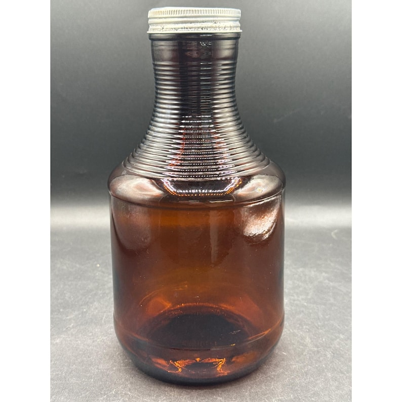 May include: A vintage brown glass bottle with a ribbed neck and a silver metal cap. The bottle has a cylindrical body and a rounded base. The glass has a slight amber tint, and the bottle appears to be empty.