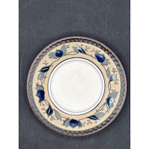 Może przedstawiać: Okrągły ceramiczny talerz z kremowym środkiem i dekoracyjną obwódką. Obwódka ma wzór niebieskich elementów kwiatowych i zielonych liści na beżowym tle. Obrzeże jest w kolorze ciemnobrązowym.