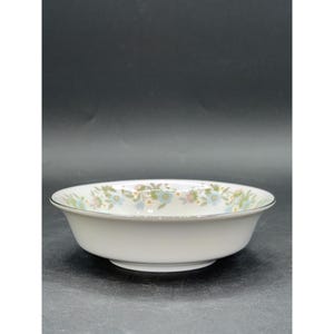 May include: A white ceramic bowl with a floral pattern around the rim. The pattern includes small blue, pink, and green flowers. The bowl has a silver trim and a slightly flared shape.