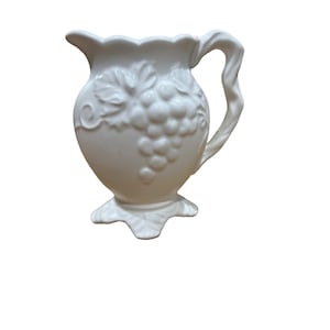 May include: White ceramic pitcher with a grapevine design. The pitcher has a footed base and a handle shaped like a vine.
