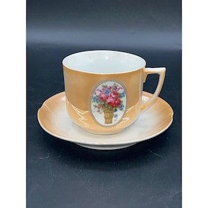 May include: A vintage teacup and saucer set with a floral design. The cup and saucer are a pale orange color with a white rim. The floral design is in a basket with pink and blue flowers.