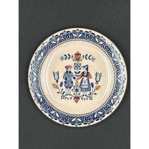 Puede incluir: Un plato redondo de cerámica con un centro color crema y un borde decorativo azul y blanco. El centro presenta una pareja con vestimenta tradicional, tomados de la mano, rodeados de motivos florales y de corazón. El plato es probablemente decorativo.