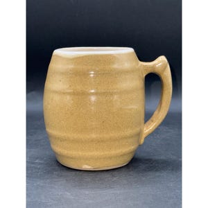 May include: A tan ceramic mug with a handle. The mug has horizontal ridges around the body and a white rim. The mug is designed for beverages.