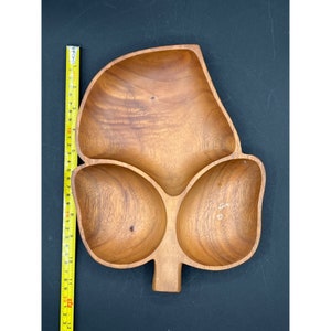 Puede incluir: Una bandeja de madera con forma de hoja con tres compartimentos y un tallo. La bandeja es de madera de color marrón claro y mide aproximadamente 33 cm de largo. La bandeja está vacía y se encuentra sobre una superficie negra.