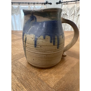 Puede incluir: Una jarra de cerámica hecha a mano con un esmalte marrón, azul y verde. La jarra tiene un cuerpo ancho y redondo y una asa.