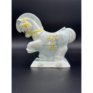 May include: A ceramic rocking horse figurine in a light blue color with yellow accents. The horse has a detailed mane and tail, and is mounted on a white base with wheels. The figurine is a decorative item.
