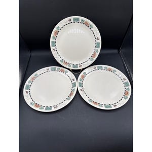 May include: Three white ceramic plates with a decorative border featuring bees, flowers, and geometric shapes in green, orange, and black. The plates are arranged on a dark surface.