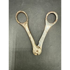 Może przedstawiać: Vintage metalowy obcinacz do knotów z dwoma okrągłymi uchwytami na palce i zawiasem. Narzędzie posiada wyblakłe, srebrzyste wykończenie z widocznymi śladami brązowego utlenienia i rdzy wzdłuż uchwytów oraz na okrągłych pierścieniach.