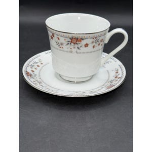 May include: A white teacup and saucer set featuring a floral design in red, orange, and grey. The cup and saucer have a silver trim. The teacup has a curved handle. The set is likely made of porcelain or a similar ceramic material.