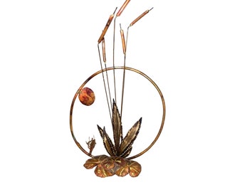 Op de afbeelding: Een wandsculptuur van koperen metaal met een gestileerde afbeelding van riet en bladeren. De sculptuur is rond en heeft een rustieke, aardse esthetiek.