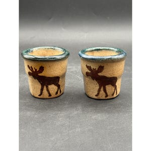 Puede incluir: Dos pequeños vasos de chupito de cerámica rústica. Cada vaso presenta una silueta de alce marrón sobre un fondo beige. Los bordes están acentuados con un esmalte de color turquesa. Los vasos tienen un aspecto artesanal, natural y terroso.