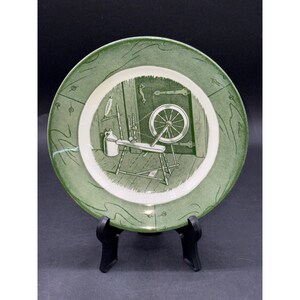 Può includere: Piatto decorativo verde e bianco con un'illustrazione dettagliata di un filatoio all'interno di una stanza. Il piatto ha un bordo verde con un motivo ondulato. L'immagine centrale è in tonalità di verde e bianco.