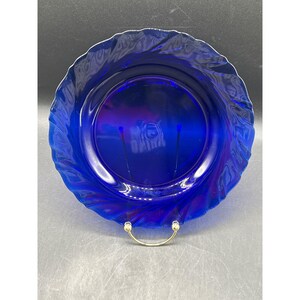 May include: A vibrant cobalt blue glass plate with a swirling, textured rim. The plate has a circular shape and is displayed on a gold-colored stand. The plate's center has the word "OMIRA" printed on it.
