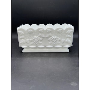 May include: A white rectangular dish with a scalloped rim and a raised grape and leaf design. The dish has a pedestal base and is made of opaque glass. The design is repeated on all four sides of the dish.