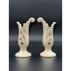 Puede incluir: Dos jarrones de cerámica blanquecina con un diseño de hojas estilizadas y una parte superior acampanada. Los jarrones tienen una base de pedestal y un remolino decorativo. El fondo es gris oscuro.