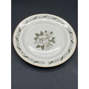 May include: A white ceramic dinner plate with a gold rim and a floral design featuring white flowers and green leaves.