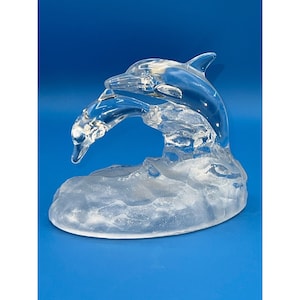 Puede incluir: Una escultura de vidrio transparente de dos delfines saltando fuera del agua. Los delfines están colocados en una pose juguetona, con un delfín saltando sobre el otro. La escultura está colocada sobre una base de vidrio transparente que se asemeja a una ola.