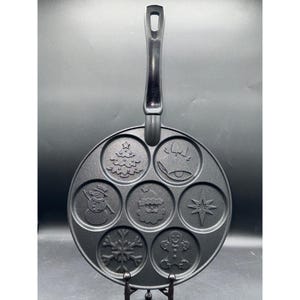 Könnte beinhalten: Eine schwarze, antihaftbeschichtete Weihnachtspfanne mit sieben kreisförmigen Formen. Zu den Designs gehören ein Weihnachtsbaum, eine Glocke, ein Schneemann, der Weihnachtsmann, ein Stern, ein Schneeflocke und ein Lebkuchenmann. Die Pfanne hat einen langen, strukturierten Griff.
