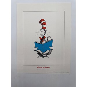Puede incluir: Una impresión con una ilustración en blanco y negro del Gato con Sombrero, que lleva un sombrero de rayas rojas y blancas y una pajarita roja, leyendo un libro azul. El texto "The Cat in the Hat" está impreso en rojo bajo la ilustración.