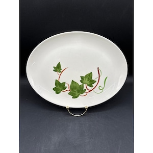 May include: Oval, off-white serving platter with a green ivy leaf design. The leaves are a vibrant green with brown and green stems. The platter is displayed on a gold-coloured metal stand against a dark background.