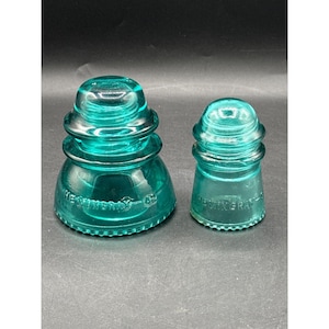 May include: Two teal glass insulators with a stepped, rounded profile. The larger insulator features the embossed text "HEMINGRAY-42" and the smaller one displays "HEMINGRAY" on the base. Both items have a smooth, translucent finish.