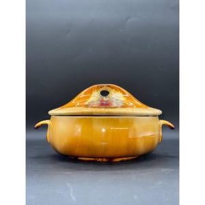 Op de afbeelding: Een vintage keramische soepterrine met een deksel. De terrine heeft een warme, amberkleurige kleur met een glanzende afwerking. Het deksel heeft een unieke vorm met een handvat en een decoratief ontwerp. De terrine heeft twee zijhandvatten.